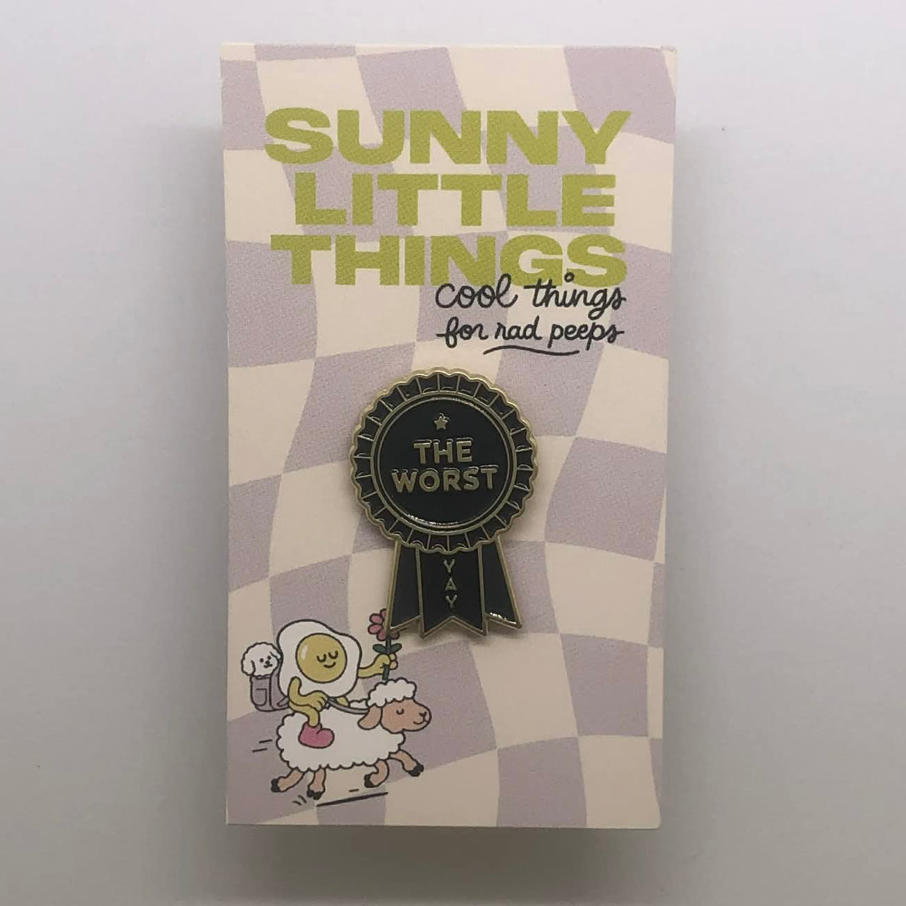 Sunny Little Things badge packaging with a badge featuring 'The Worst' text on a checkered background.